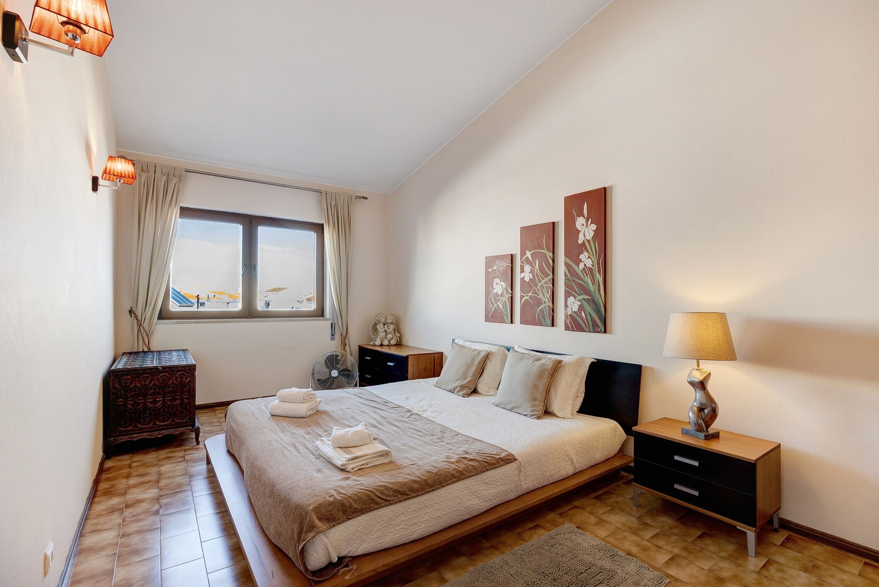 A bedroom with a large bed, a bedside table, a nightstand, a window with curtains, and wall art.