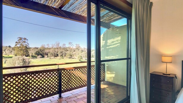 A balcony with a view of a grassy field and trees, a wooden fence, a window with a view of a building, and a lamp on a dresser.