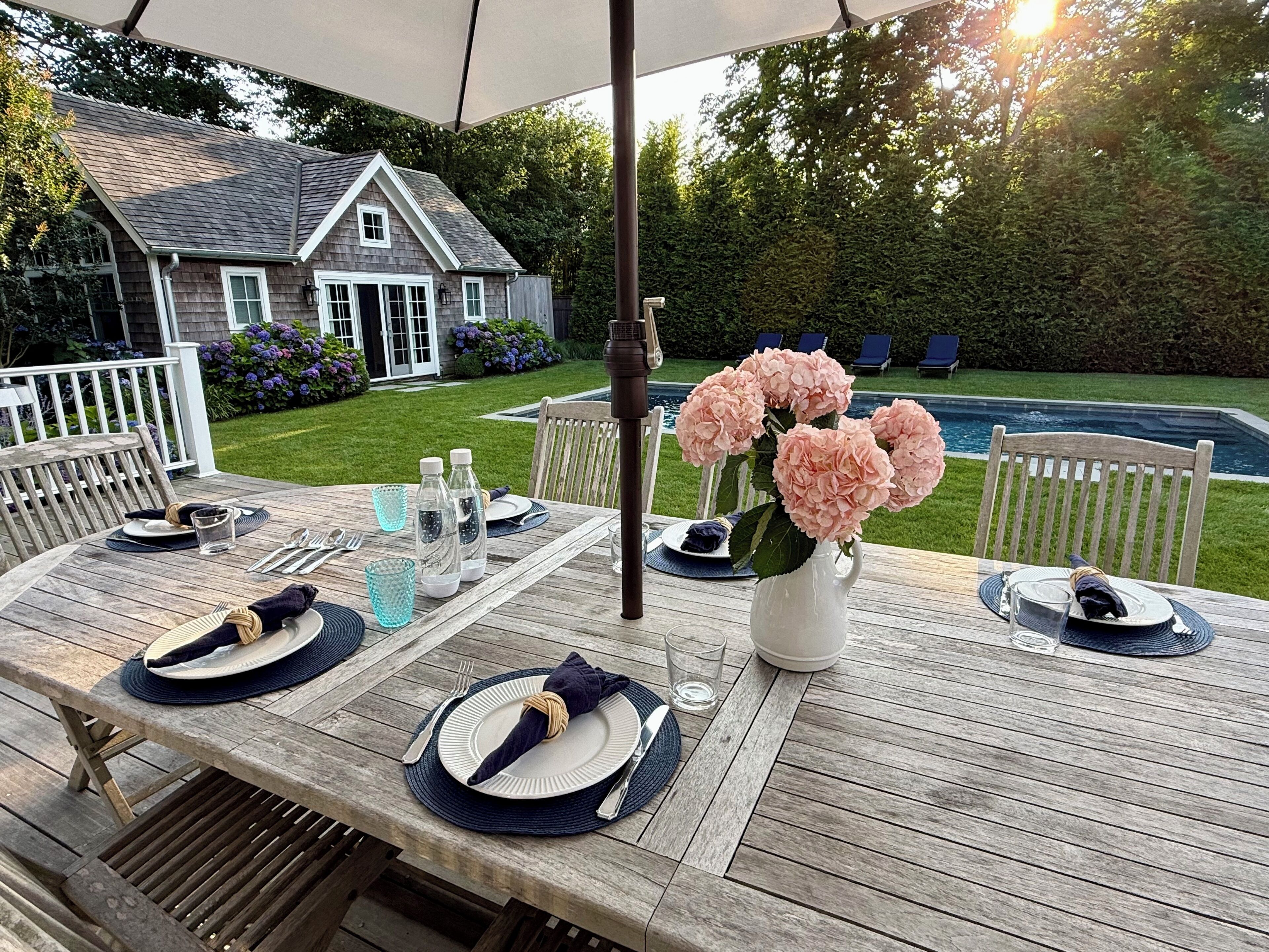 An outdoor dining area with a wooden table set for four, surrounded by a white fence, a house, and a swimming pool in the background.