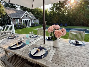 Un área de comedor al aire libre con una mesa de madera dispuesta para cuatro personas, rodeada por una cerca blanca, una casa y una piscina al fondo.
