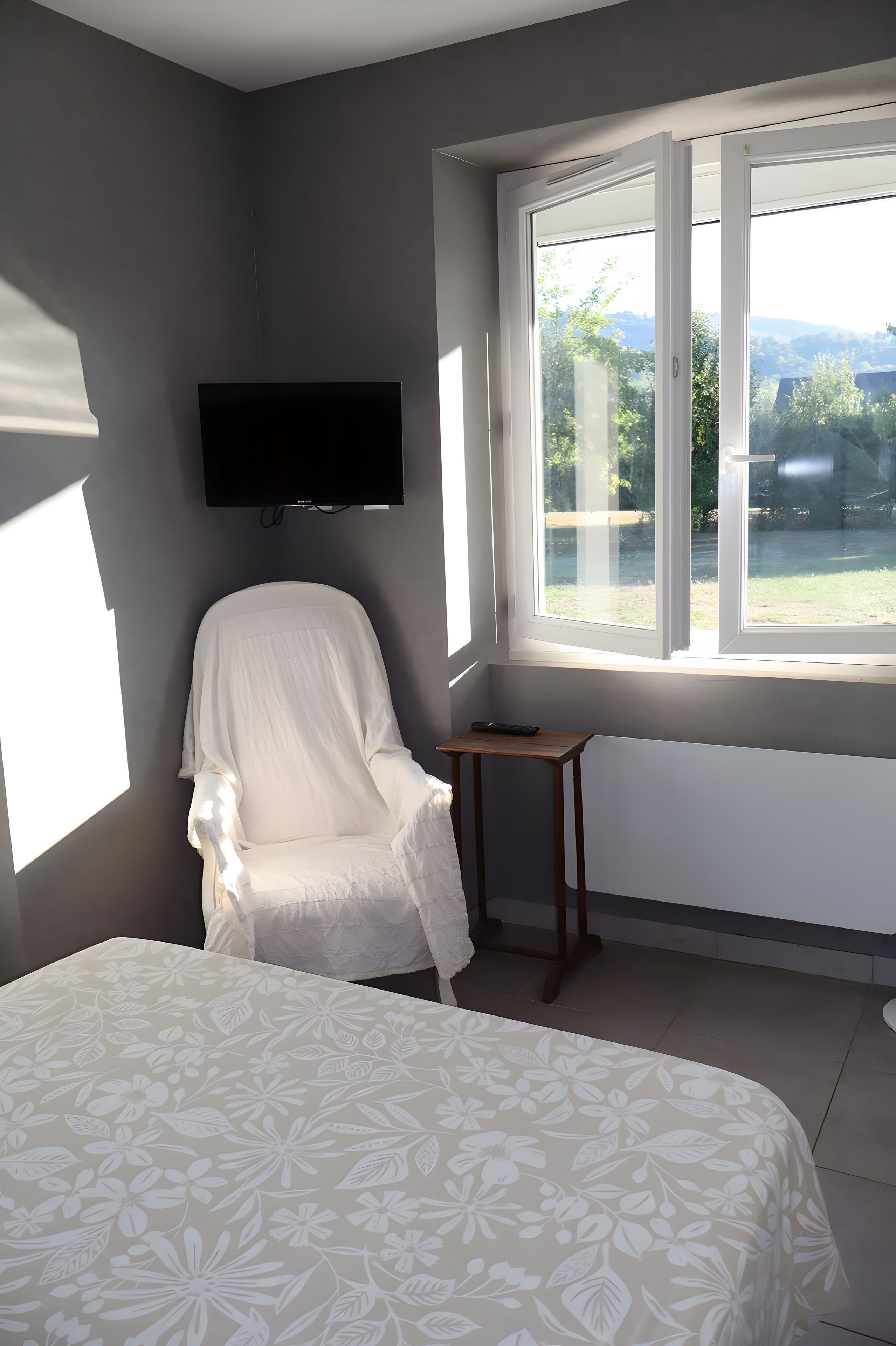 Een slaapkamer met een bed, een stoel, een klein tafeltje, een aan de muur bevestigde televisie en een raam met uitzicht op groen.