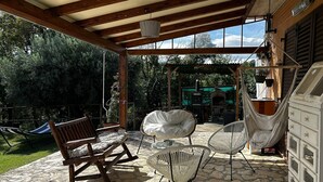 A covered patio with stone flooring, wooden furniture, and a view of a garden and outdoor kitchen.
