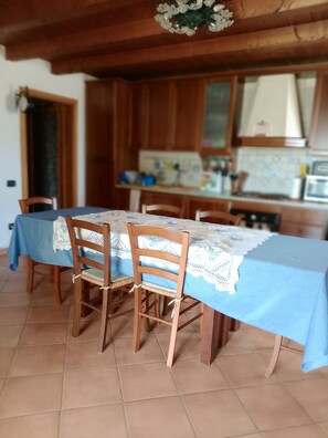 A dining area with a table covered in a blue tablecloth, wooden chairs, and a kitchen in the background.