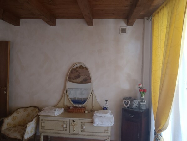 A room with a wooden ceiling, a white dresser with a mirror, a chair, and a small table with a vase of flowers.