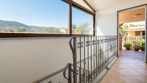 Un balcón con barandilla de metal, vistas a las montañas y un edificio con plantas en macetas.