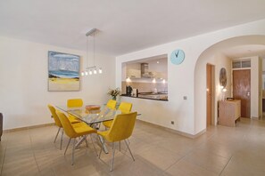 Un comedor moderno con una mesa de vidrio y sillas amarillas, una cocina con un reloj, y un cuadro en la pared.