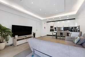 Un salon moderne avec un téléviseur à écran plat, un canapé gris et un coin repas avec des chaises, et une cuisine en arrière-plan.