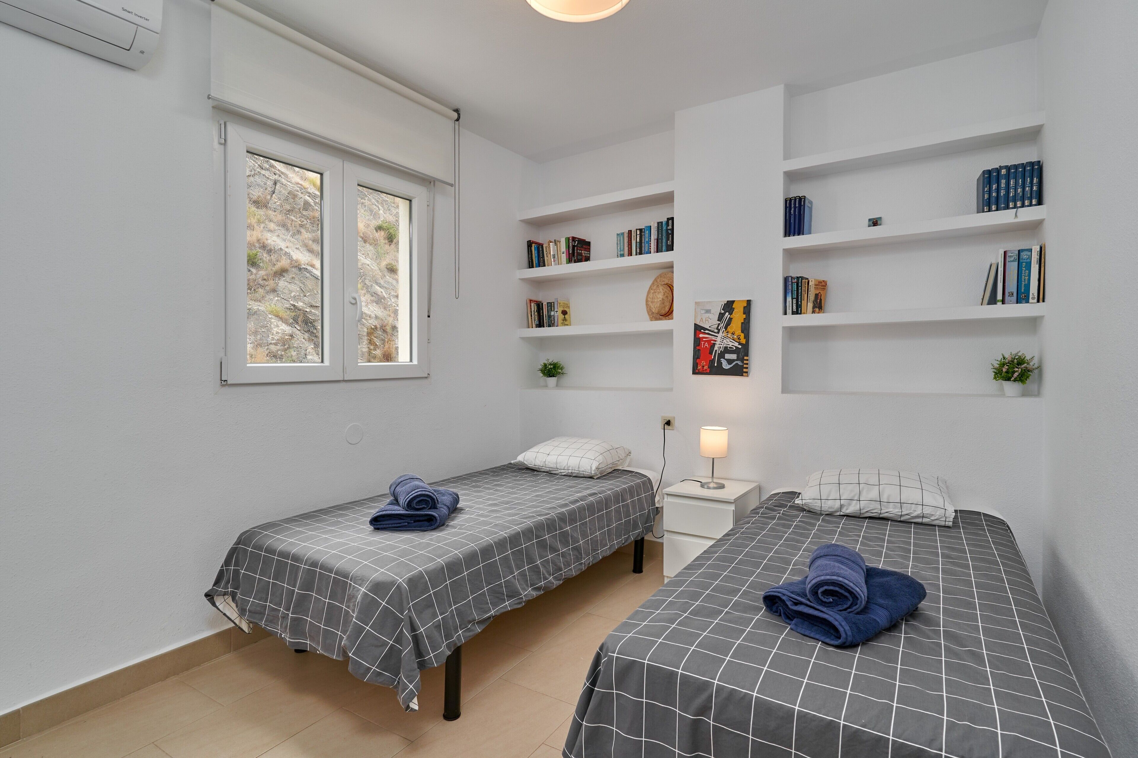 A bedroom with two beds, a bookshelf, a window, and a bedside table with a lamp.