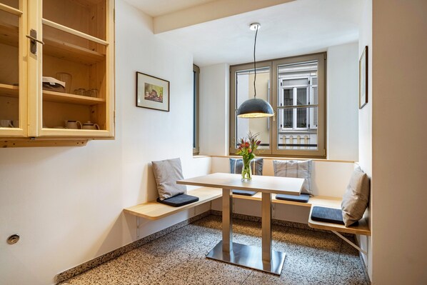 Une pièce avec un meuble en bois, une table sur laquelle est posé un vase, un banc avec des coussins, une lampe suspendue et une fenêtre avec des stores.