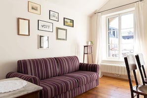 Un salon avec un canapé à rayures, une petite table recouverte d’une nappe en dentelle, une chaise, un radiateur et une fenêtre avec des rideaux.