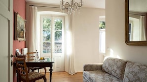 A room with a dining table, chairs, a sofa, a chandelier, and a large window with curtains. - Apartment 'Villa Bergmann Steinach Suite' with Mountain View, Shared Garden and Wi-Fi (Meran)