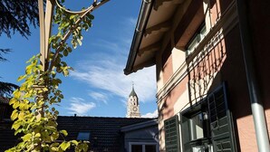 Un edificio con una torre de reloj, un balcón con estructura metálica y un ventanal con contraventanas.
