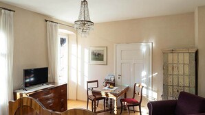 Una habitación con una cama, un escritorio con televisor, una mesa de comedor con sillas, una araña y una puerta.