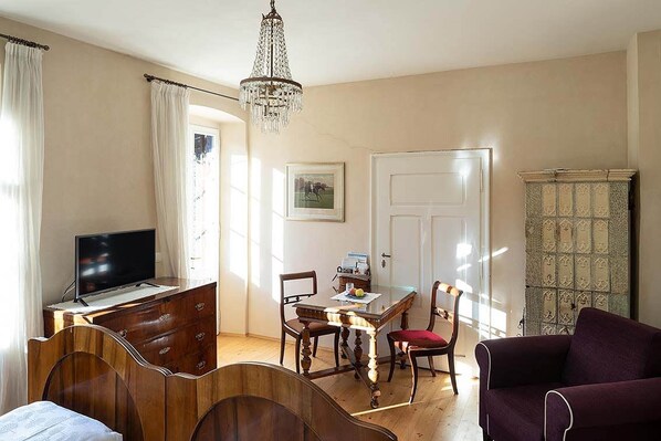 Una habitación con una cama, un escritorio con televisor, una mesa de comedor con sillas, una araña y una puerta.