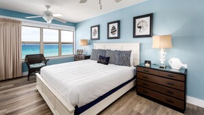 Ein Schlafzimmer mit einem großen Bett, einem Nachttisch, einer Kommode und einem Sessel. An den Wänden hängen Bilder und das Fenster bietet Blick aufs Meer.