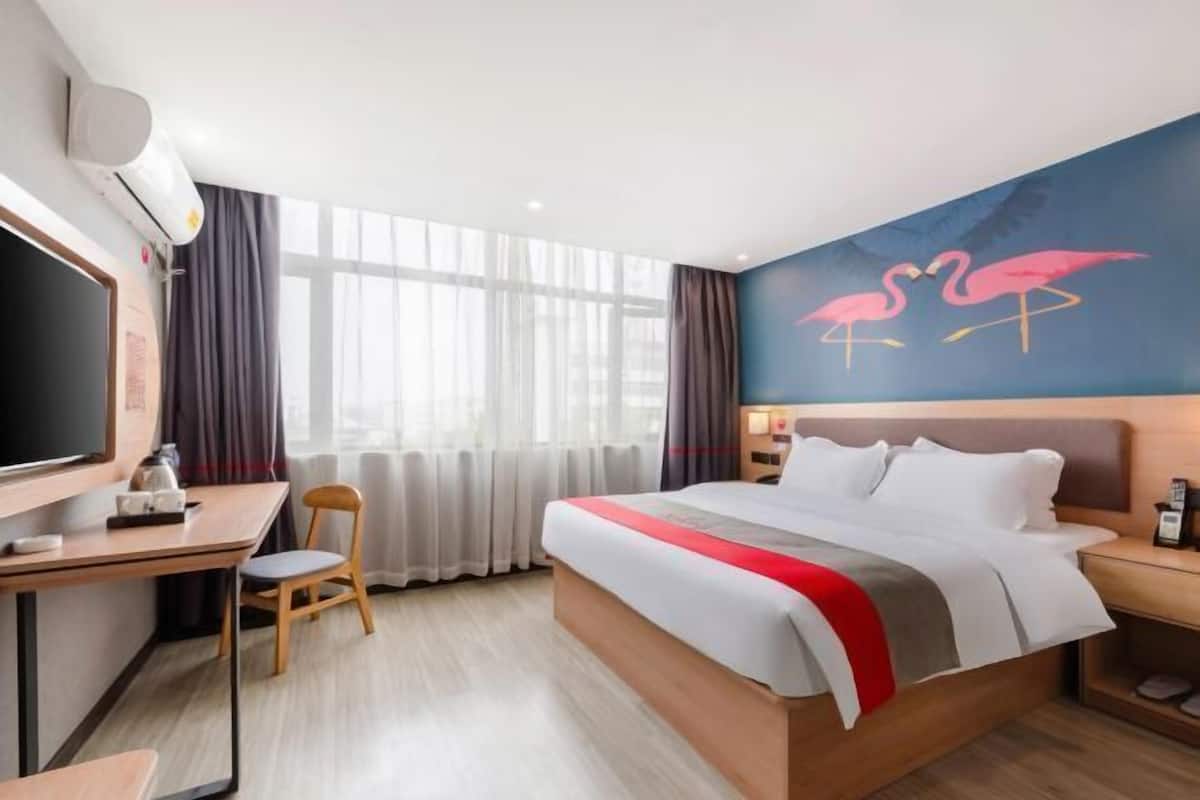 A hotel room with a bed, a desk, a chair, a TV, and a wall with a flamingo mural.