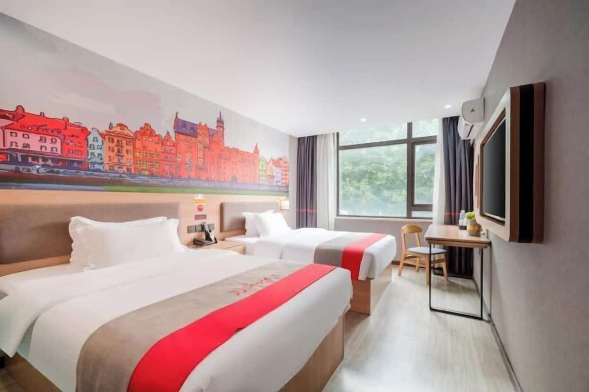 A hotel room with two beds, a desk, a chair, a TV, and a mural of a cityscape.