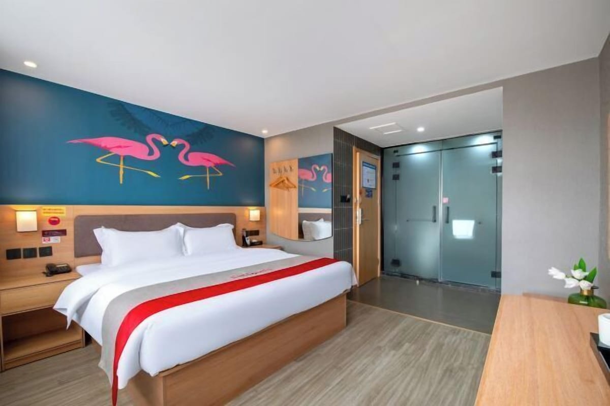 A hotel room with a large bed, a nightstand, a desk, a mirror, and a wall with a flamingo mural.