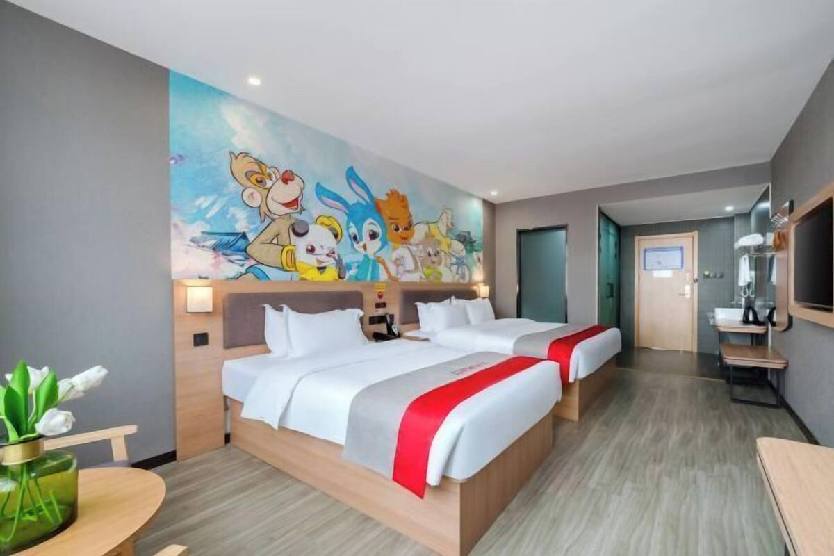 A hotel room with two beds, a mural on the wall, a small table with a vase, and a bathroom visible through an open door.