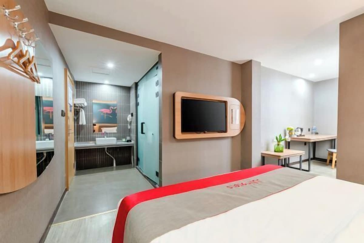 A modern hotel room with a bed, a TV mounted on the wall, a desk with a chair, and a bathroom visible through an open door.