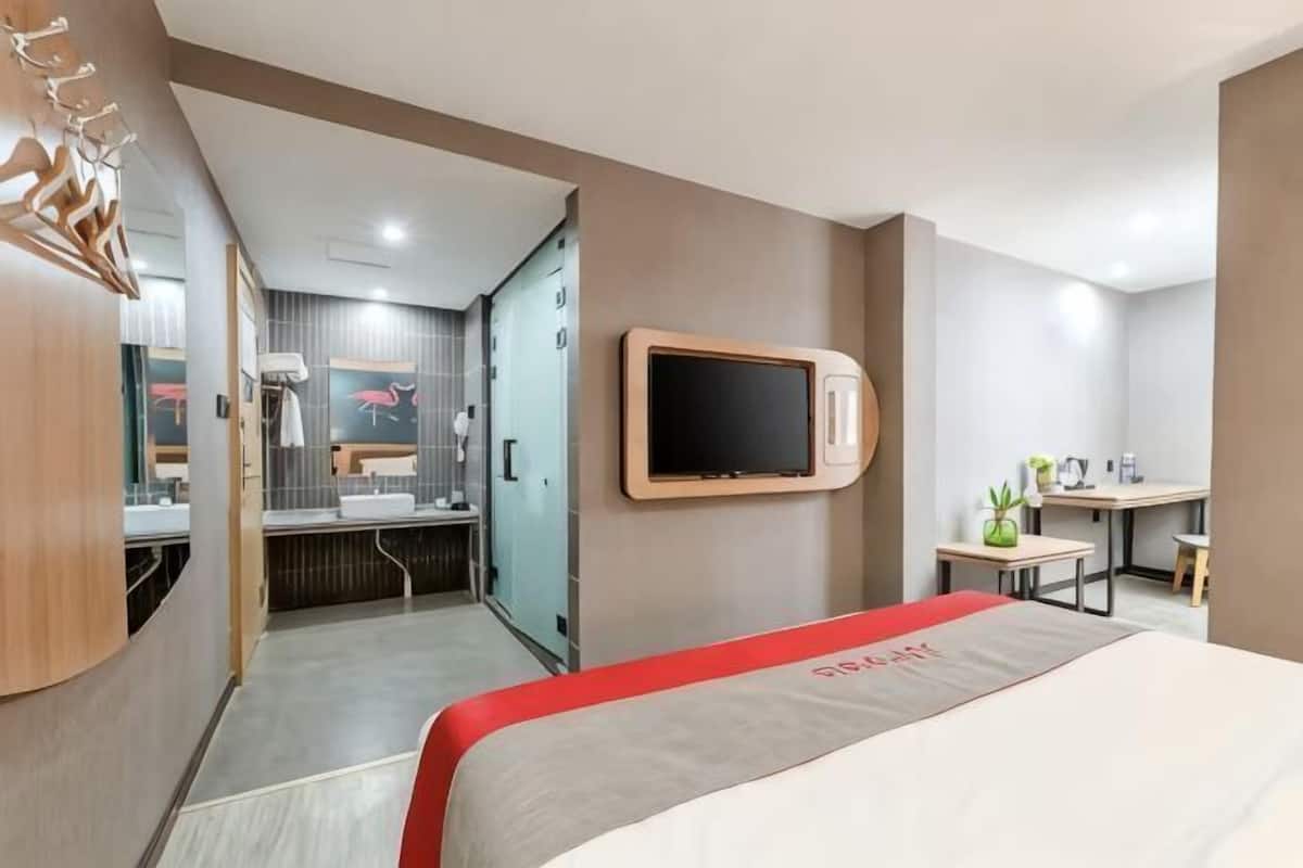 A hotel room with a bed, a TV mounted on the wall, a desk, and a small table with a plant.