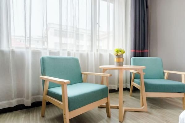 A room with two teal chairs and a small wooden table, a vase with flowers, sheer curtains, and a view of buildings through large windows. - Junyi Hotel (Huaibin Mobile Building Store) (Huaibin)