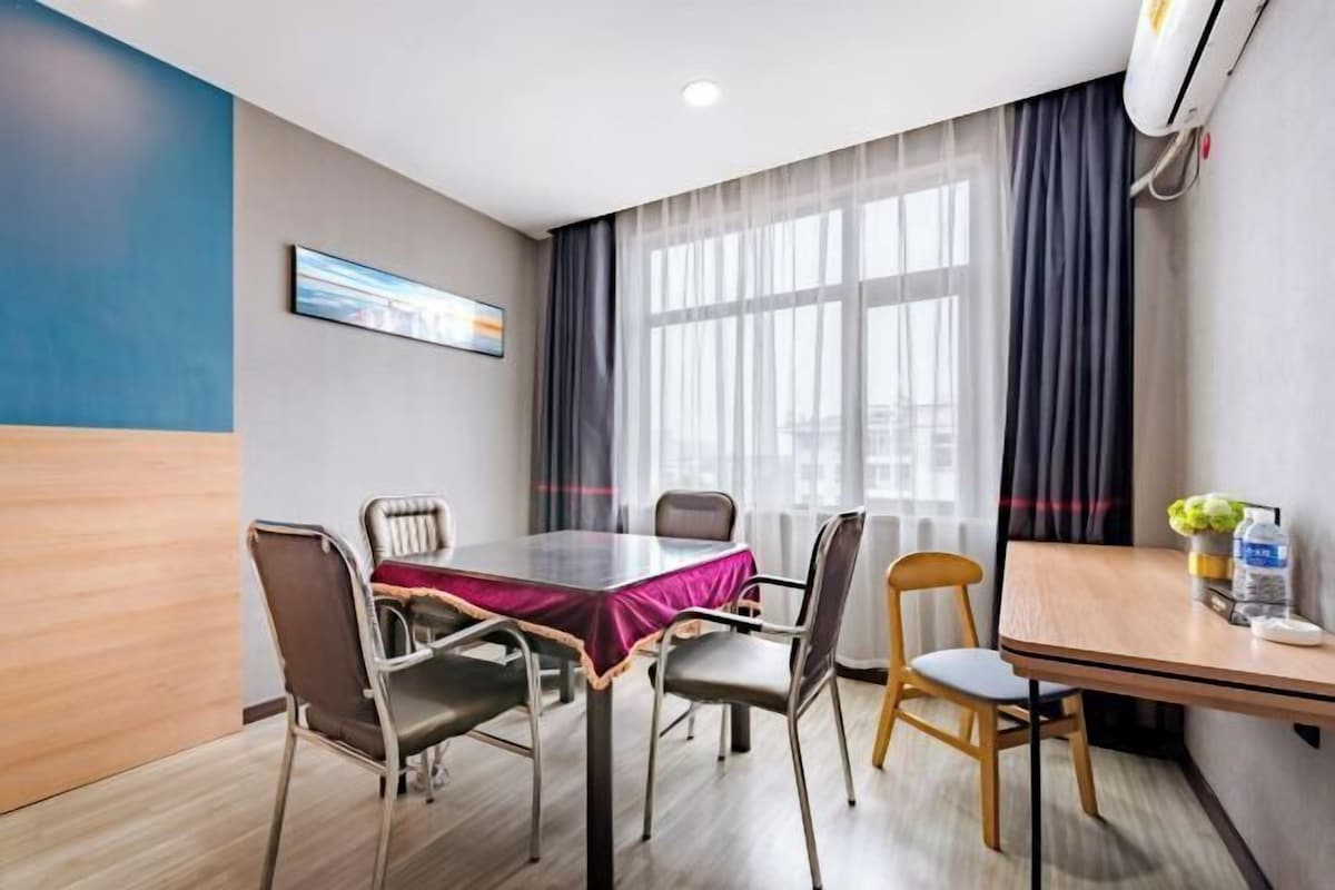 A hotel room with a dining table, chairs, a painting on the wall, a window with curtains, and an air conditioning unit.