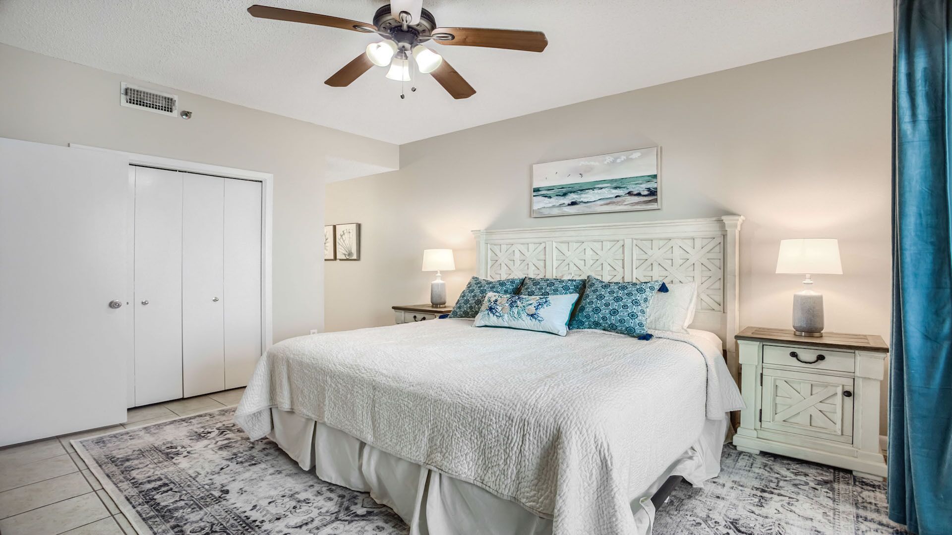A bedroom with a bed, two bedside tables with lamps, a ceiling fan, a door, and a wall-mounted painting.