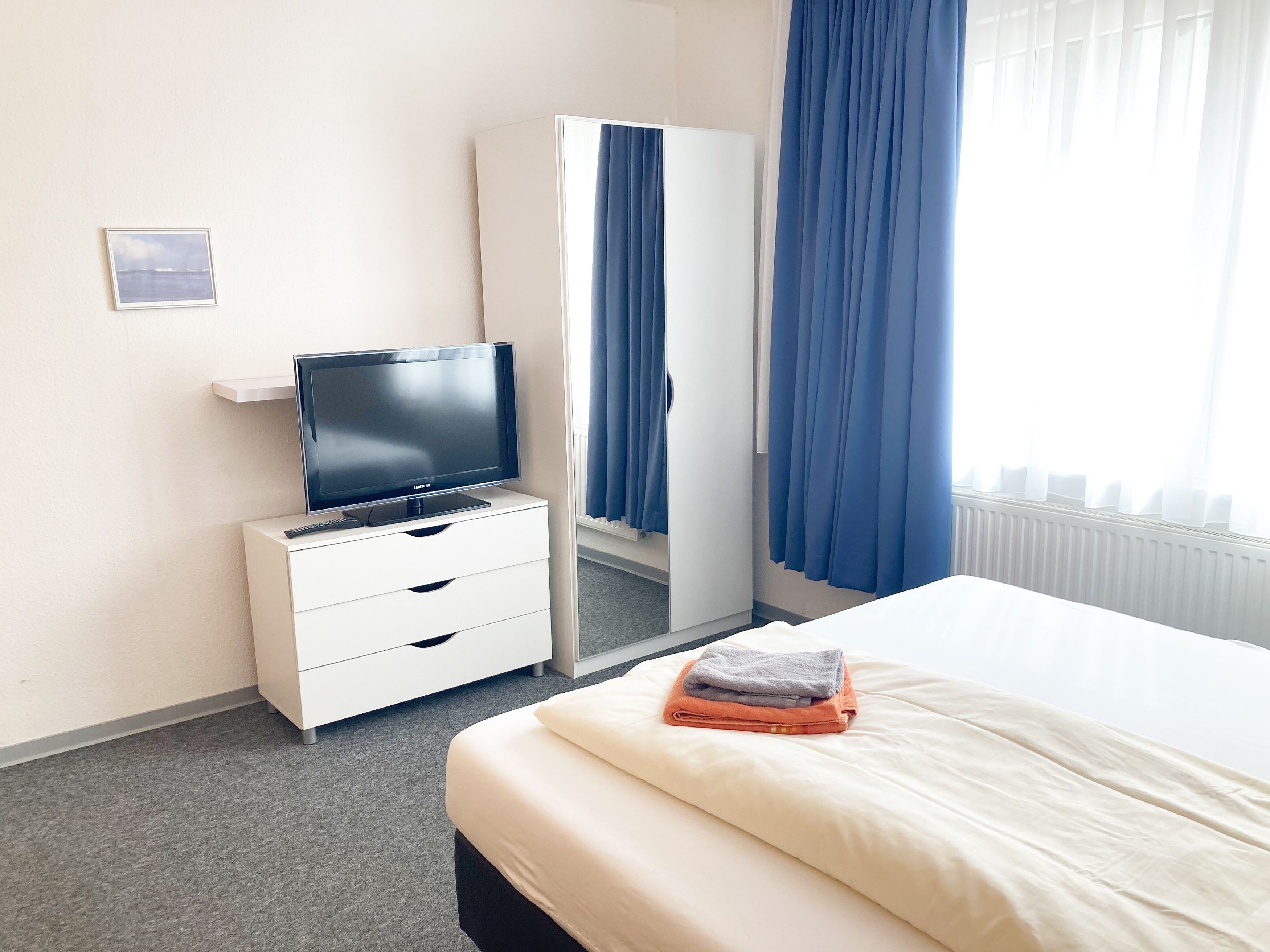 Habitación de hotel con una cama, un televisor sobre un tocador blanco, un armario y una ventana con cortinas azules.