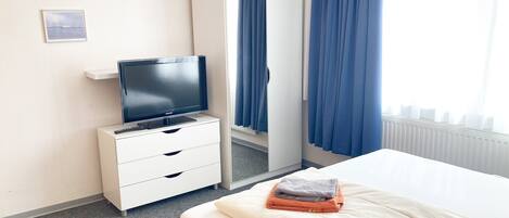 Habitación de hotel con una cama, un televisor sobre un tocador blanco, un armario y una ventana con cortinas azules.