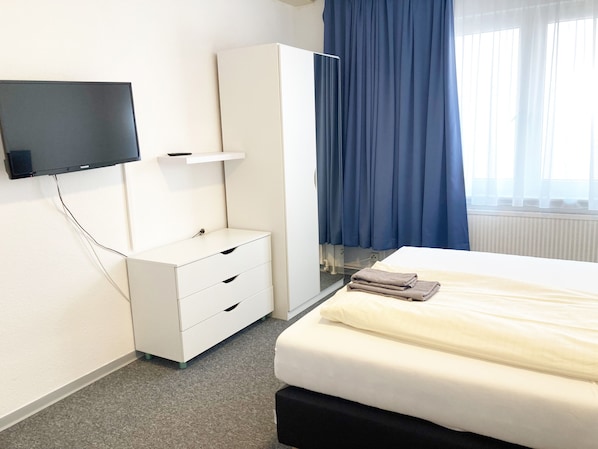 Una habitación de hotel con una cama, un televisor montado en la pared, un armario y una ventana con cortinas.