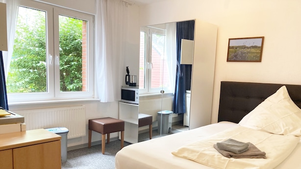 Een slaapkamer met een bed, een bureau, een stoel, een klein krukje, een kledingkast en een raam met uitzicht op groen.