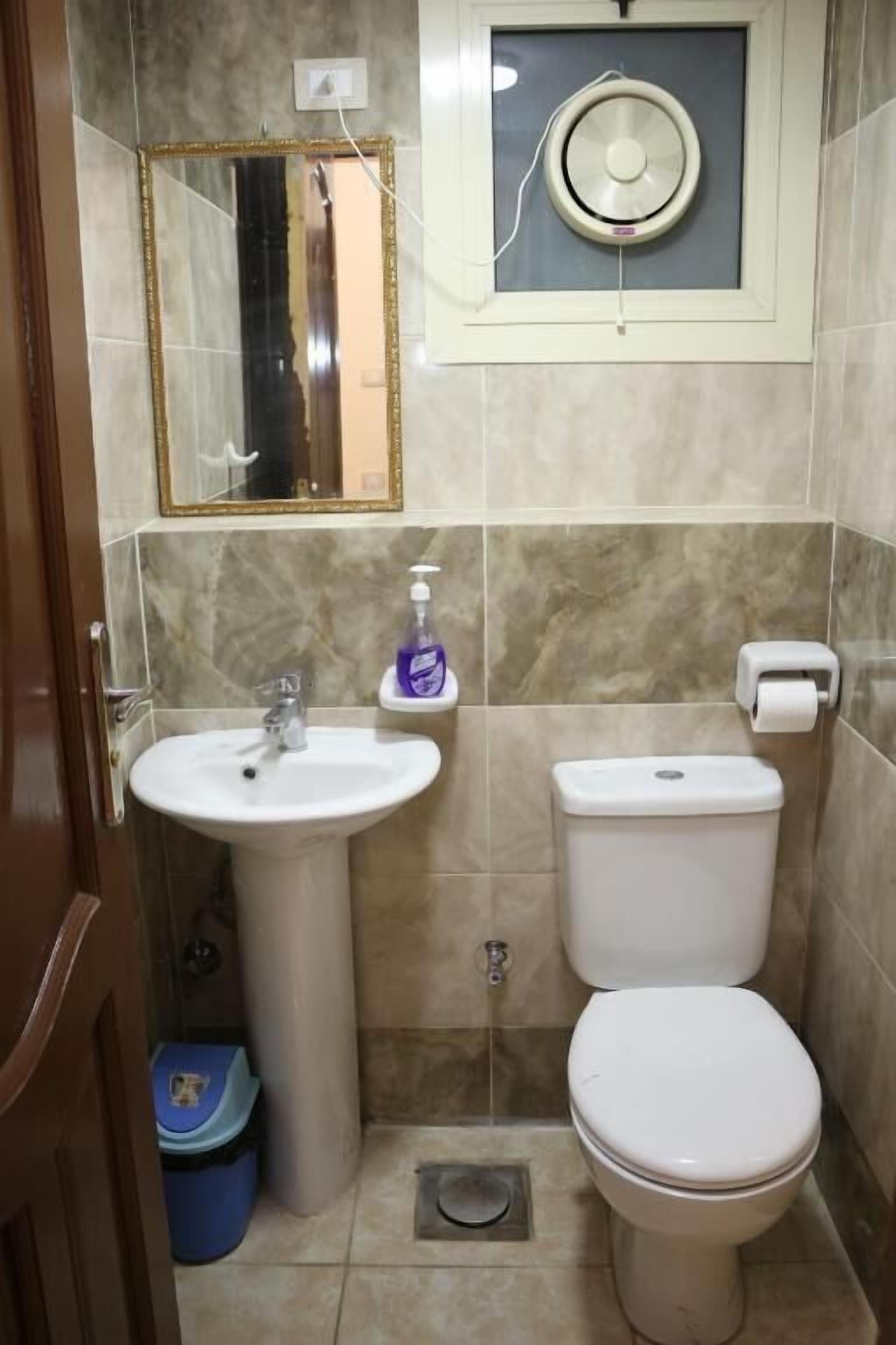 A bathroom with a sink, toilet, mirror, and a small window with a fan.