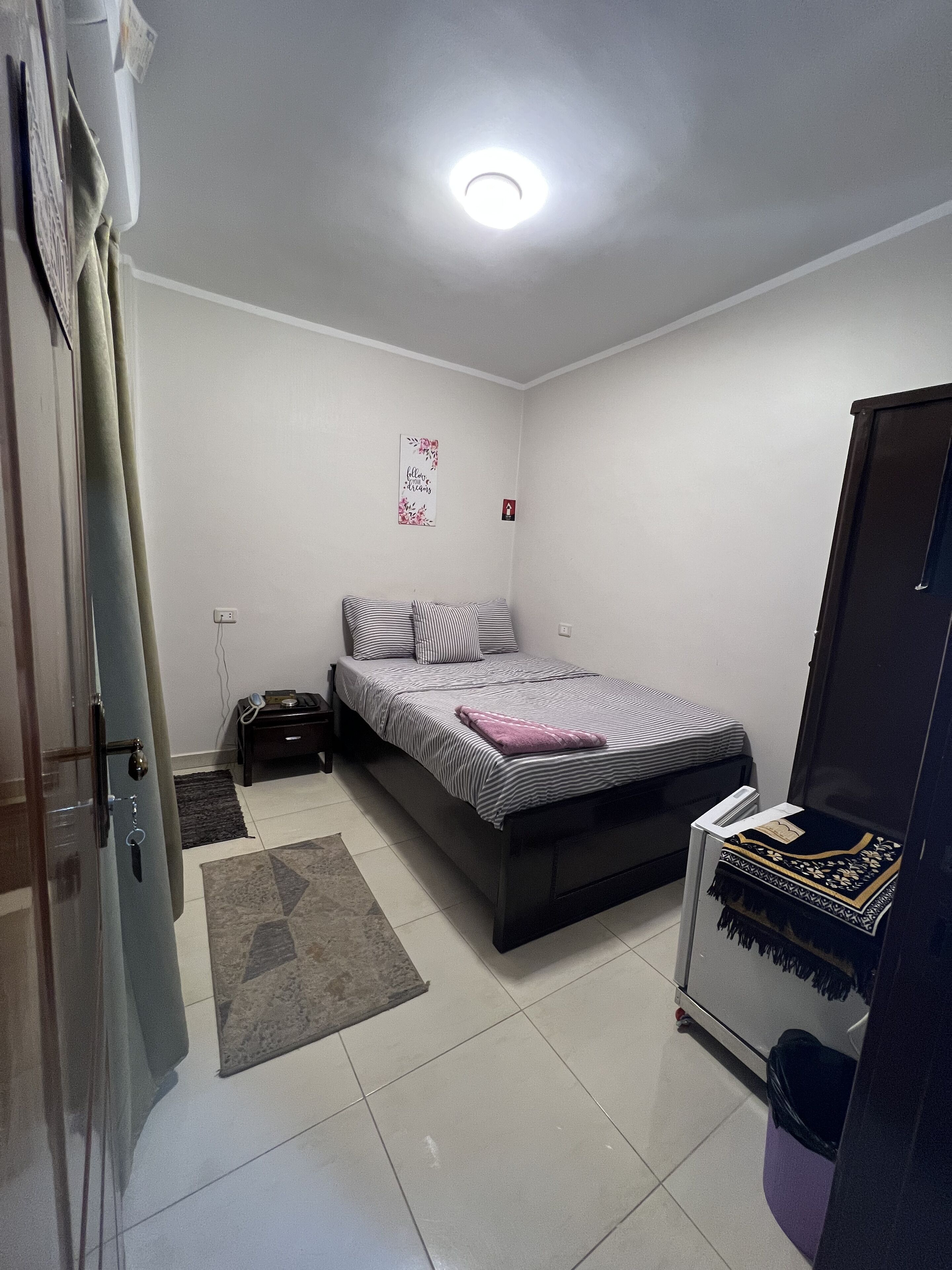 Un dormitori amb un llit, una tauleta de nit, una catifa petita, una decoració a la paret i una porta.