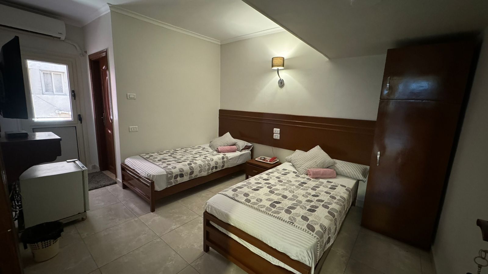 Habitación de hotel con dos camas individuales, un escritorio, un armario y una lámpara de pared.