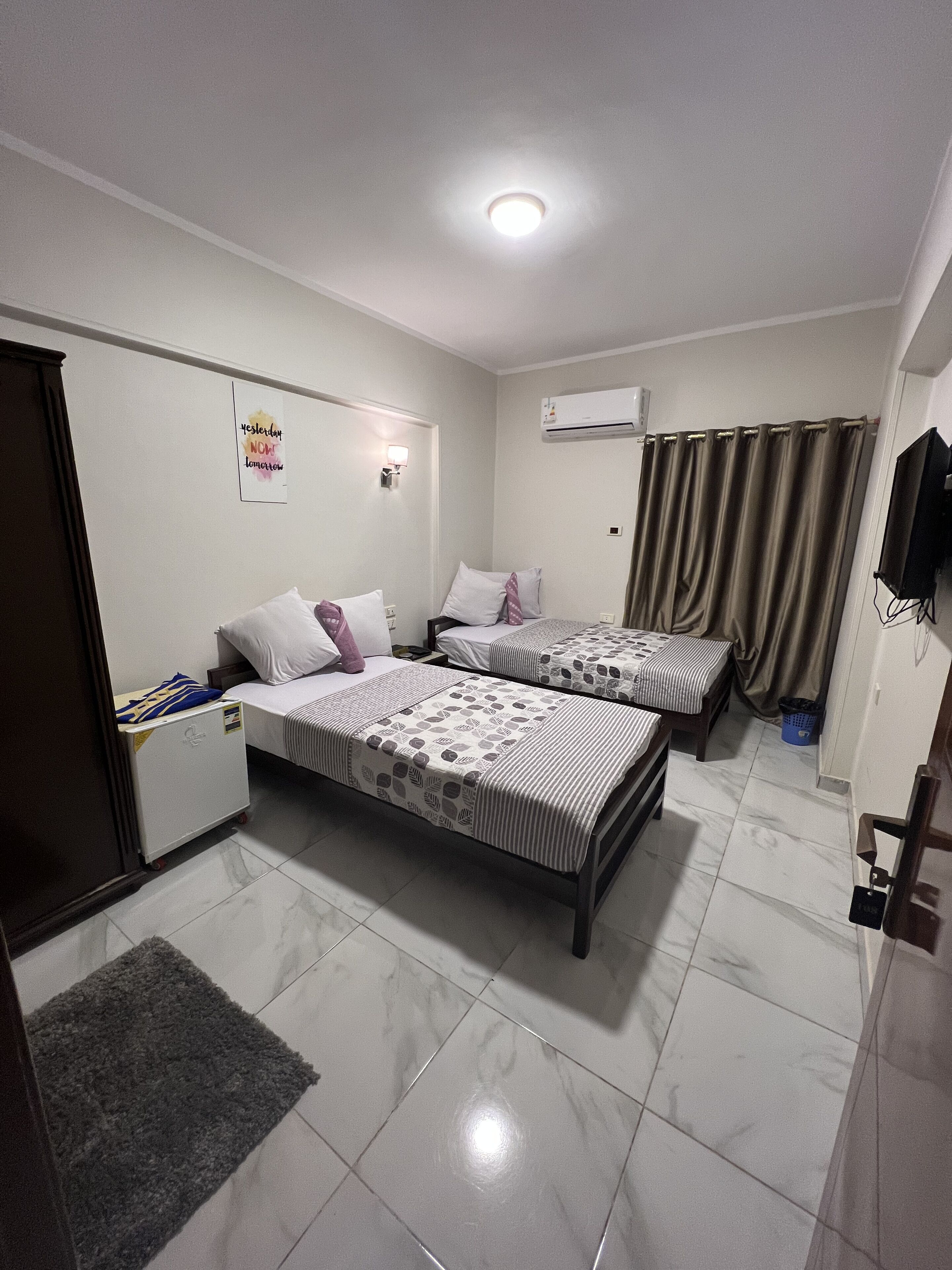 A hotel room with two beds, a small desk, a chair, a TV, an air conditioning unit, and a door.
