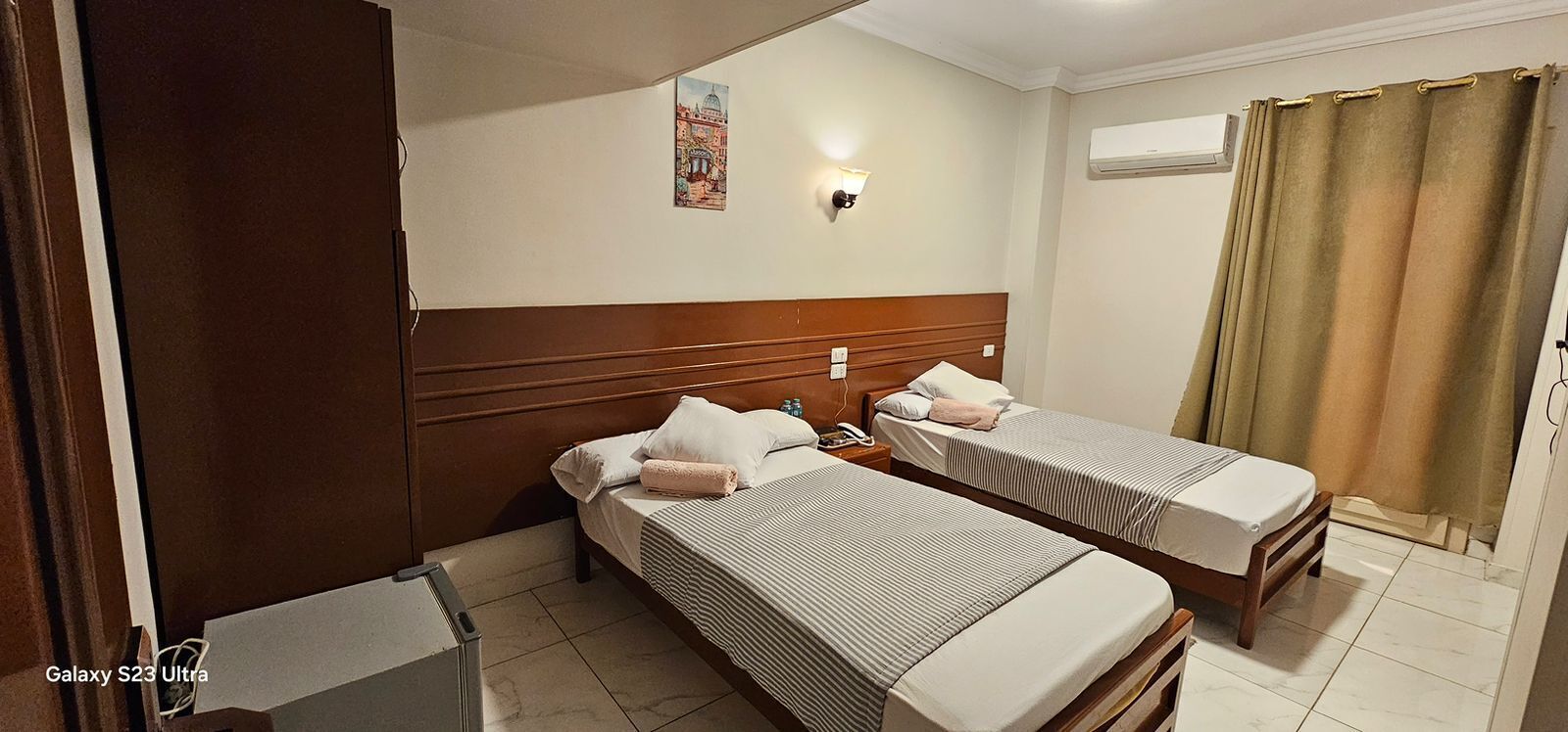 Una habitación de hotel con dos camas individuales, cabecera de madera, mesita de noche con teléfono, luz fijada en la pared y una ventana con cortinas.
