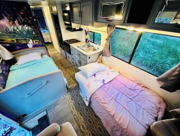 Een compact camperinterieur met twee bedden, een kitchenette en een groot raam met uitzicht op een groen landschap.