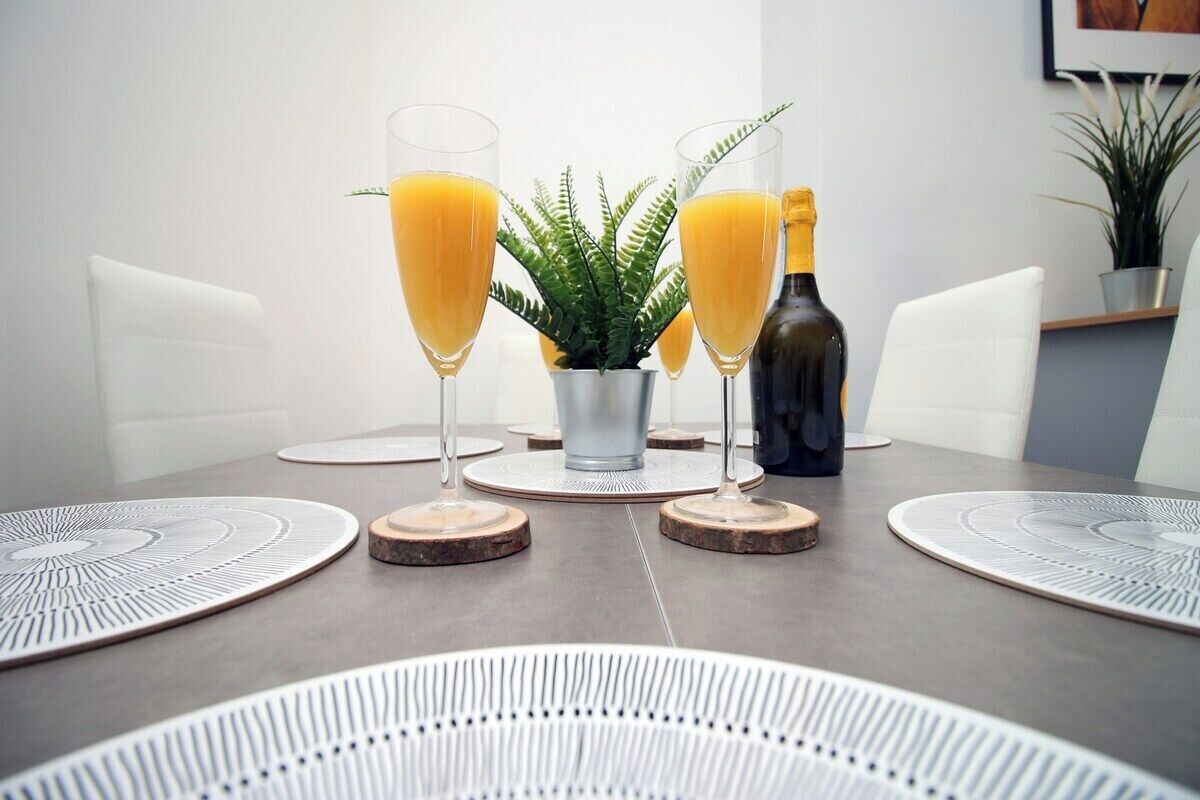 A dining table with two glasses of orange juice, a bottle of champagne, and a small potted plant.