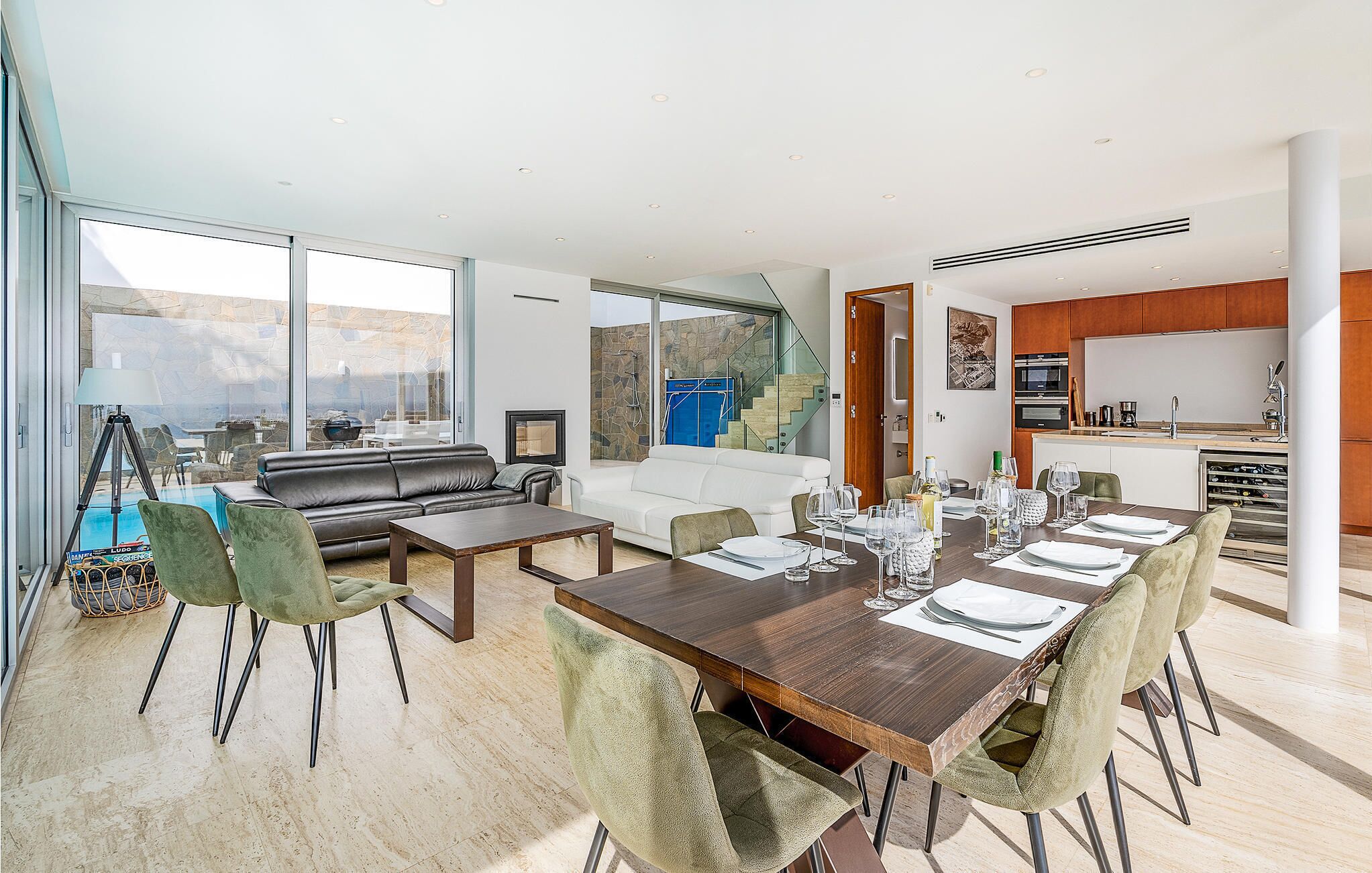 Una sala de estar moderna con comedor, ventanas amplias, chimenea y cocina visible a través de una puerta abierta.