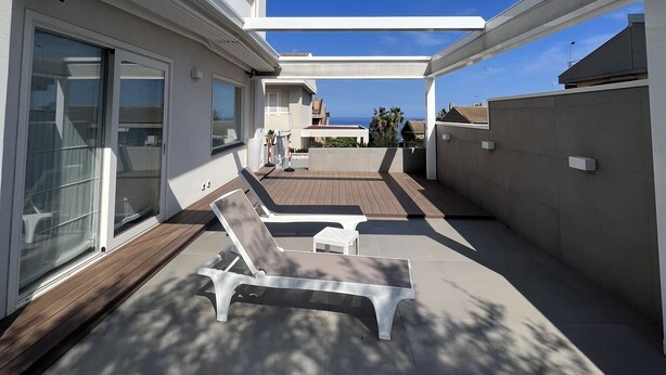 A modern outdoor patio with a view of the sea, featuring a wooden deck, lounge chairs, and a pergola.