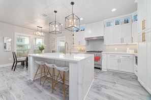 A modern kitchen with white cabinetry, a central island with stools, pendant lighting, and a stainless steel range hood. - Sunkissed Starfish: Heated Pool & Spa, beach access, Galveston (Galveston)