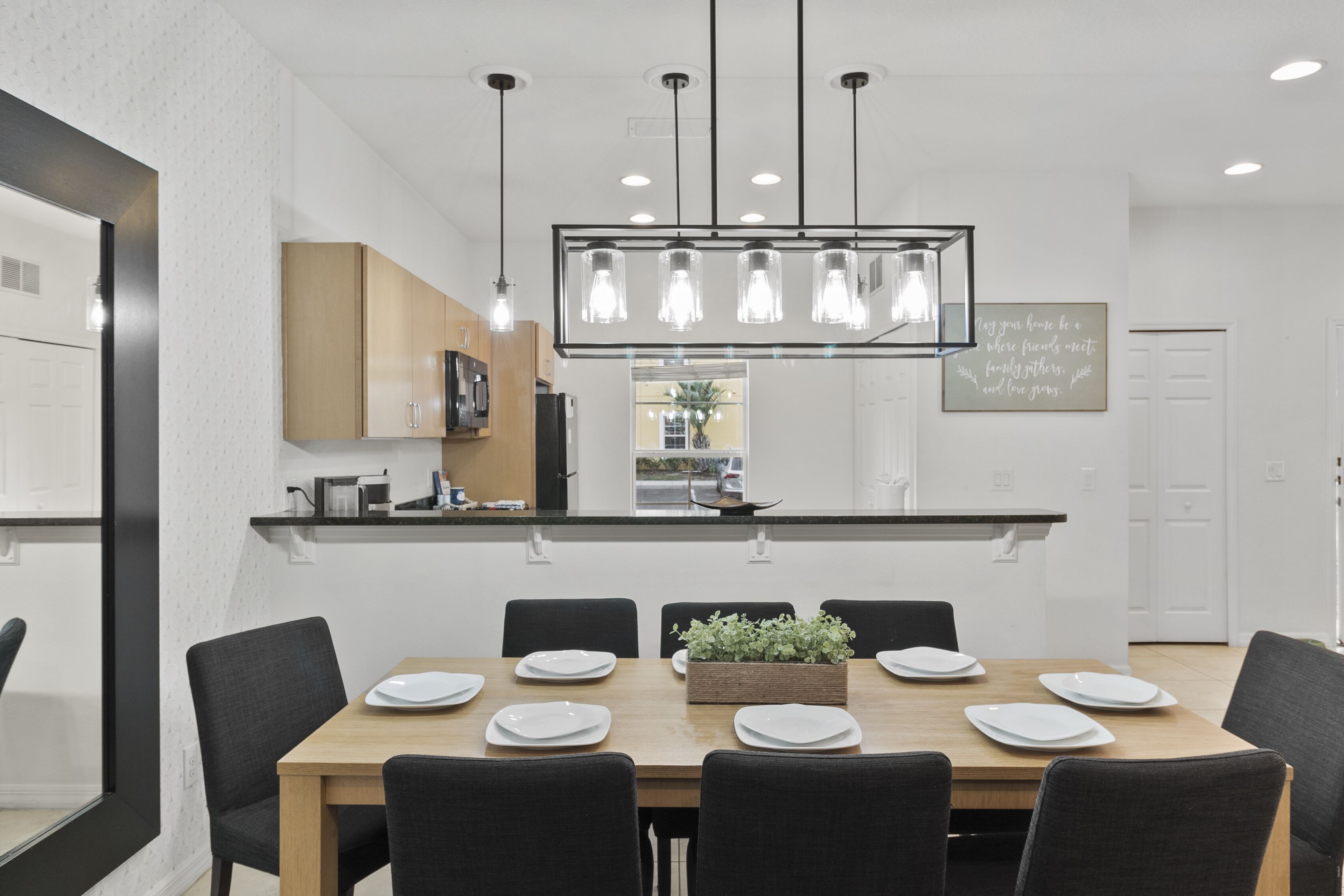 A modern dining area with a wooden table set for six, surrounded by black chairs. A hanging light fixture with multiple bulbs is above the table.