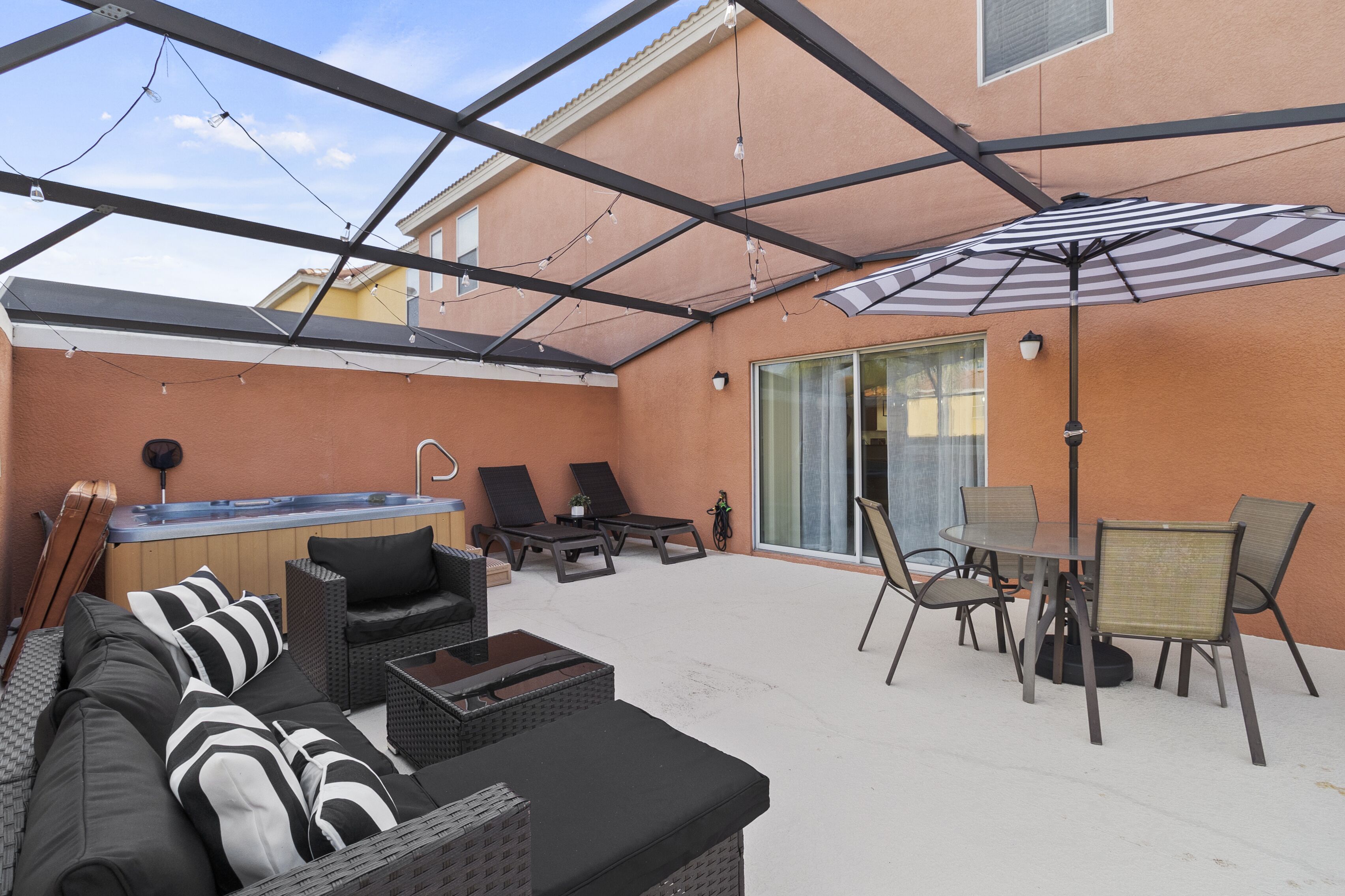 Un patio techado al aire libre con un jacuzzi, sillones reclinables, una mesa de comedor con sillas y un paraguas grande.