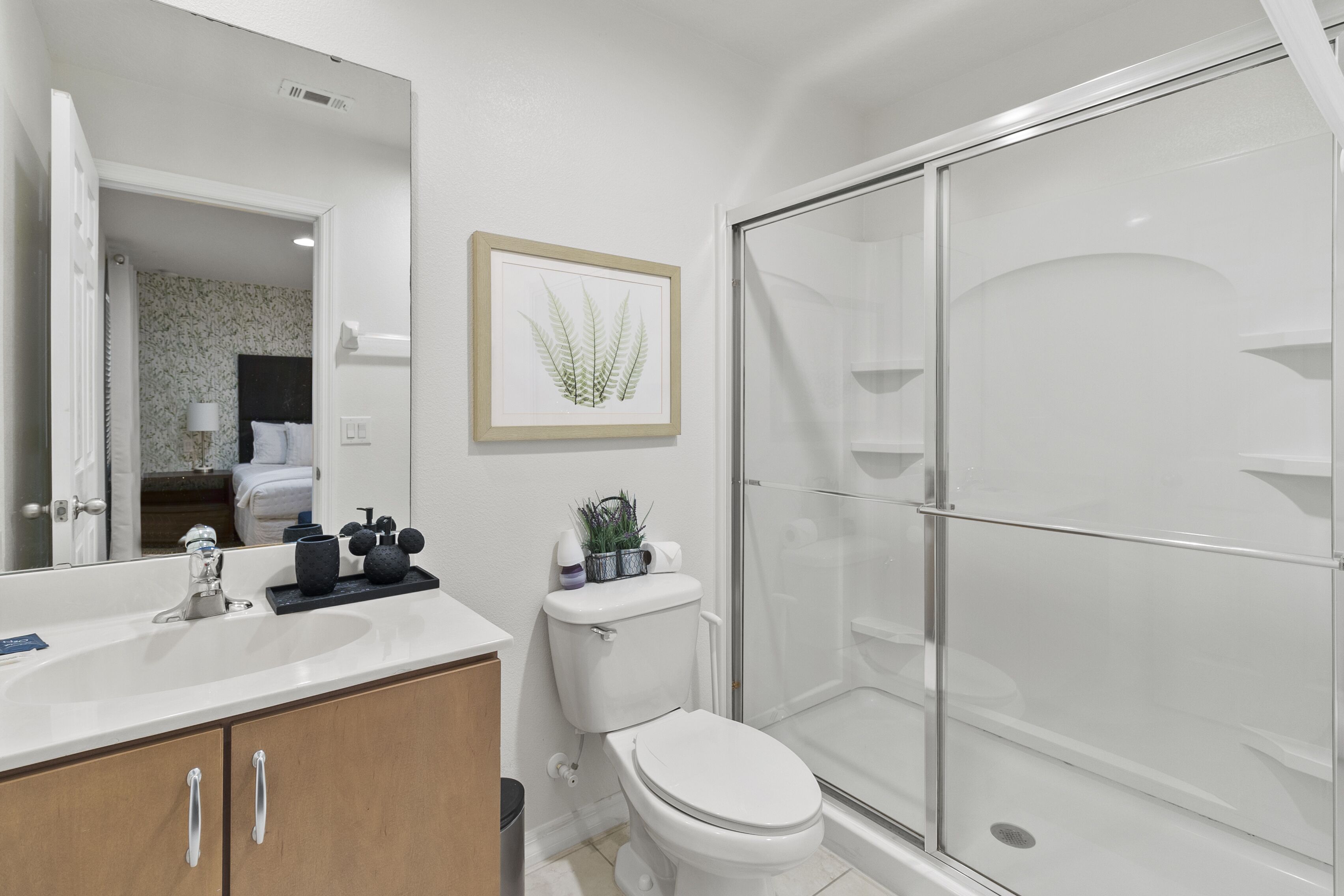 Un bagno moderno con lavandino bianco, mobile contenitore in legno, una pianta incorniciata, il water e un box doccia in vetro.