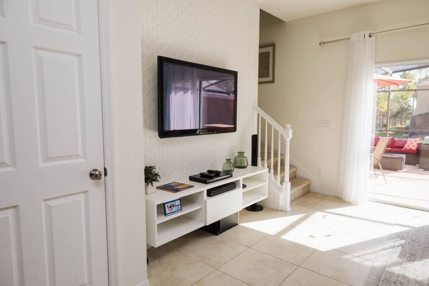 Habitación con televisor de pantalla plana montado en la pared, una consola blanca con objetos decorativos, una escalera y vistas a un patio exterior.