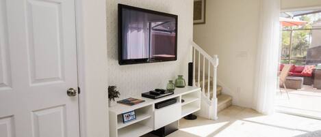 Habitación con televisor de pantalla plana montado en la pared, una consola blanca con objetos decorativos, una escalera y vistas a un patio exterior.