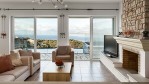 A spacious living room with a large TV, comfortable seating, a fireplace, and a scenic ocean view through large windows. - The Hillside Villas, Stunning Views & Private Pool (Corfu)