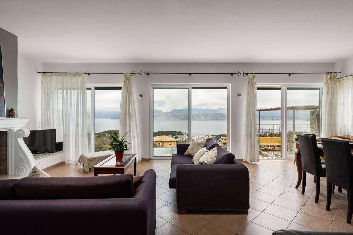 A spacious living room with a fireplace, a sofa, a dining table with chairs, and large sliding glass doors offering a view of the sea and mountains.