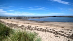 A sandy beach with a wooden pier extending into the water, a clear sky, and a distant lighthouse. - The Anchorage - sea views and beach on doorstep (Amble)